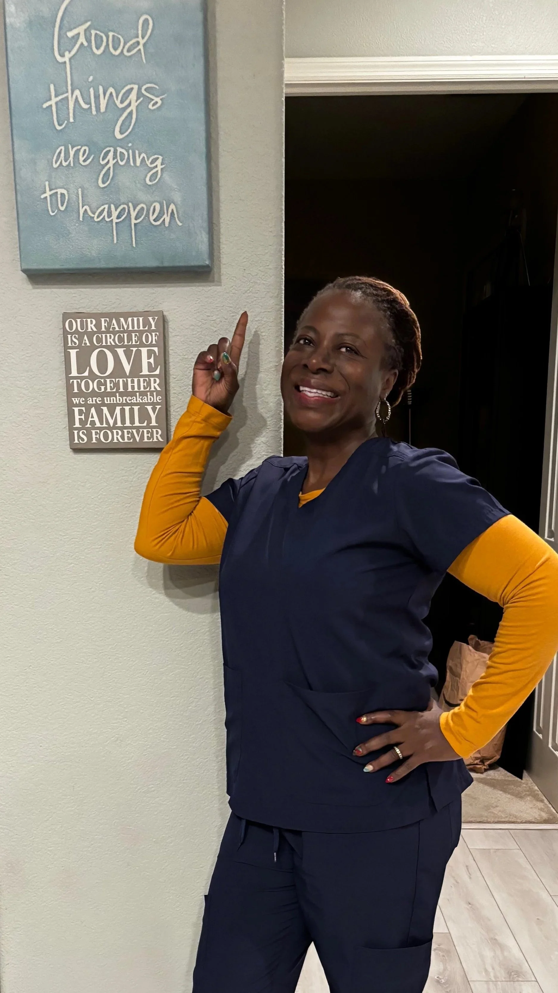 A smiling woman with braids, wearing navy medical scrubs with yellow sleeves, pointing at wall decorations and standing in a room with light-colored walls and wooden flooring.