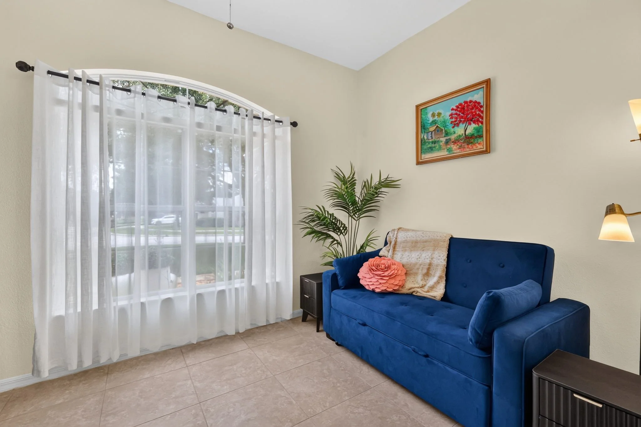 Living room corner with cream-colored walls, large window with white sheer curtains, blue velvet sofa with pink pillow and beige blanket, a wall painting of a house, trees, and a red tree, small black side table, and a floor lamp.