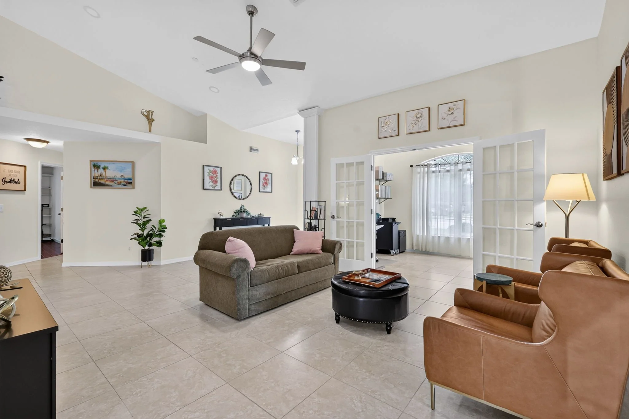 Living room with beige tiled floor, beige walls, a ceiling fan, and a variety of furniture including a beige sofa with pink pillows, a brown leather armchair, and a small black ottoman. There are framed pictures and decorative items on the walls, a t