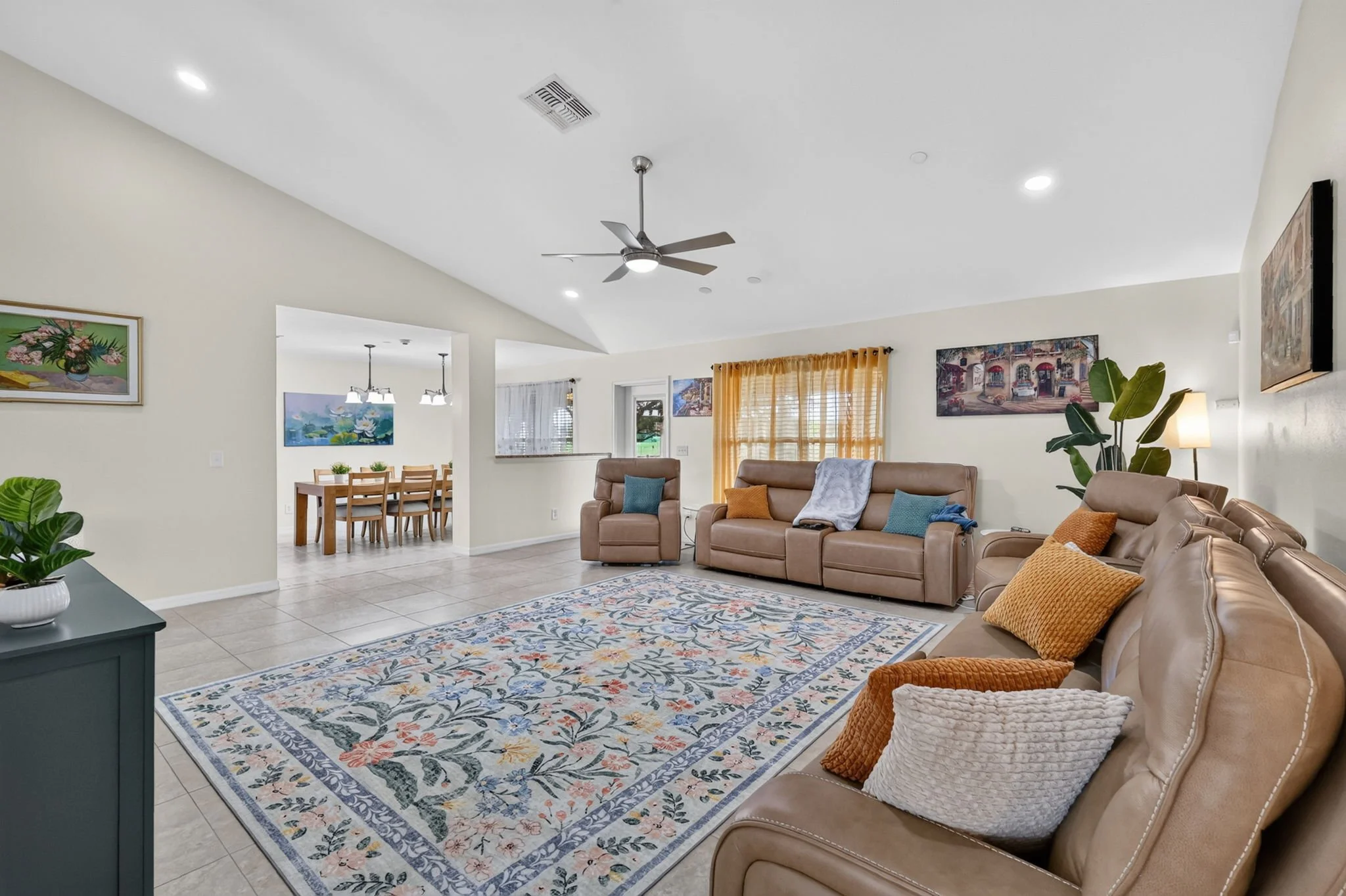 A living room with beige sofas, colorful throw pillows, a patterned area rug, a large potted plant, artwork on the walls, and a ceiling fan. There's a dining area with a wooden table and chairs visible through an open doorway.