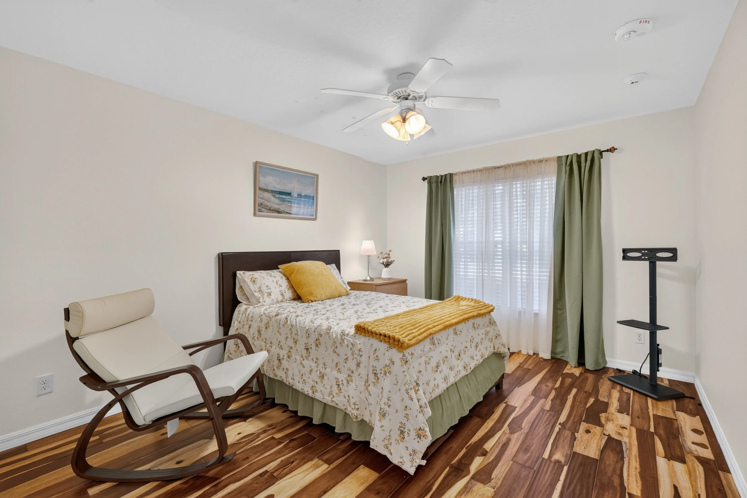A bedroom with a bed, side table, and window with green curtains. A ceiling fan with lights is turned on, and there's a framed picture on the wall. An armchair is in the corner, and a TV stand is on the right.