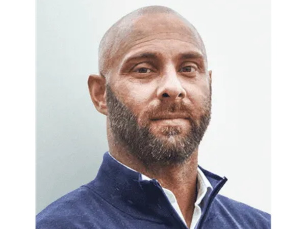 A middle-aged man with a bald head and beard, wearing a blue zip-up jacket and white shirt, looking at the camera against a plain background.