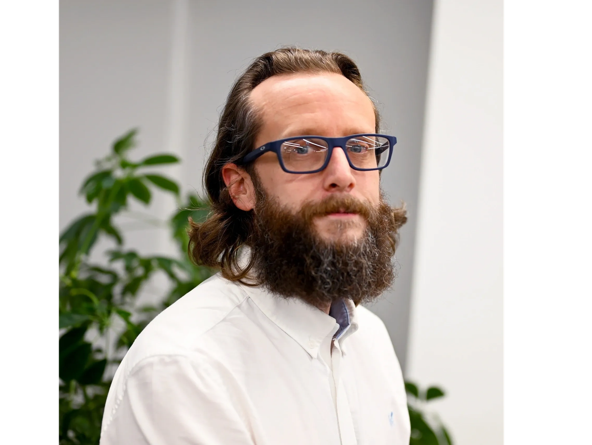 A man with long brown hair, a full beard, glasses, wearing a white shirt, standing indoors with a plant in the background.
