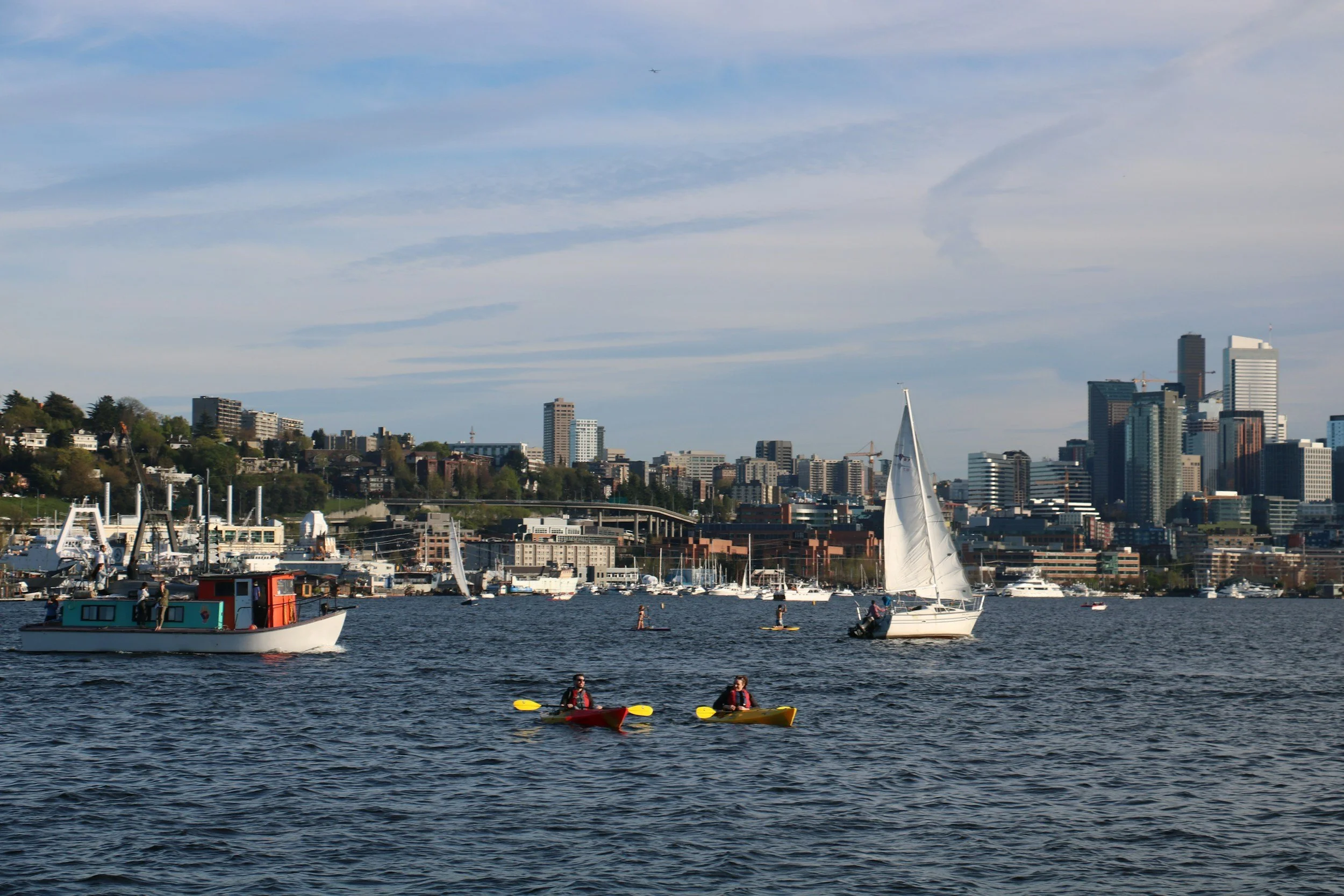 A city skyline with high-rise buildings and cranes visible across a body of water. There are boats and people kayaking on the water.