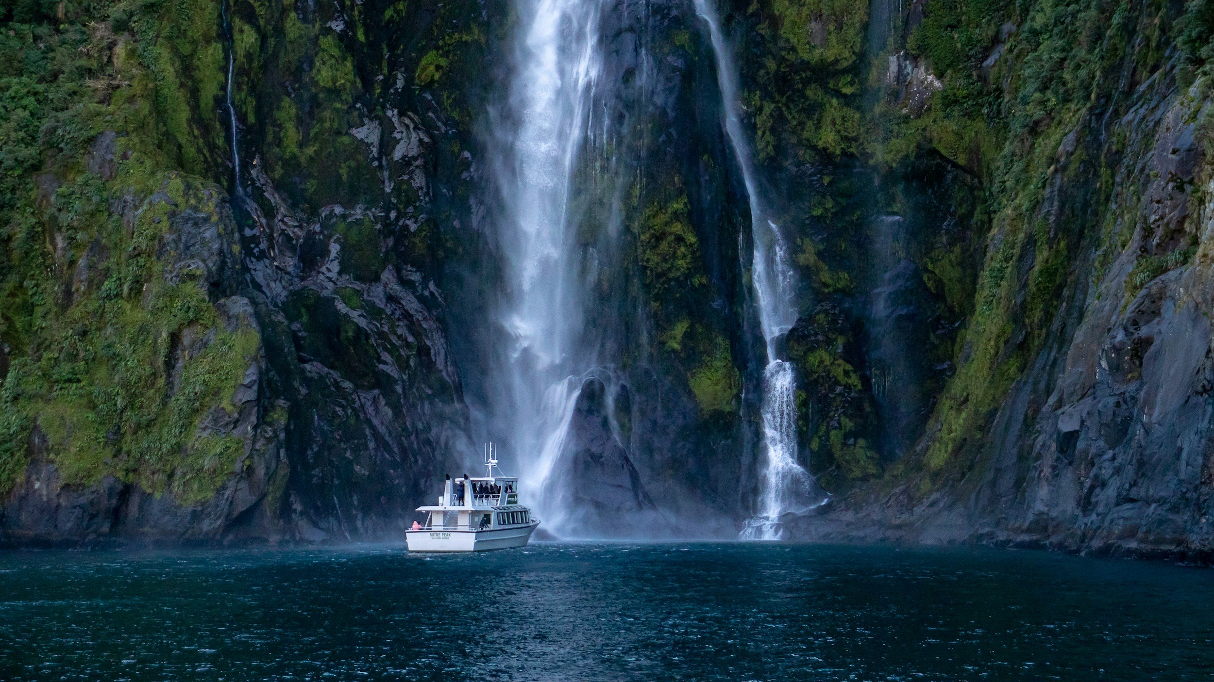 A boat navigating through a narrow body of water with a large waterfall cascading down moss-covered cliffs in the background.