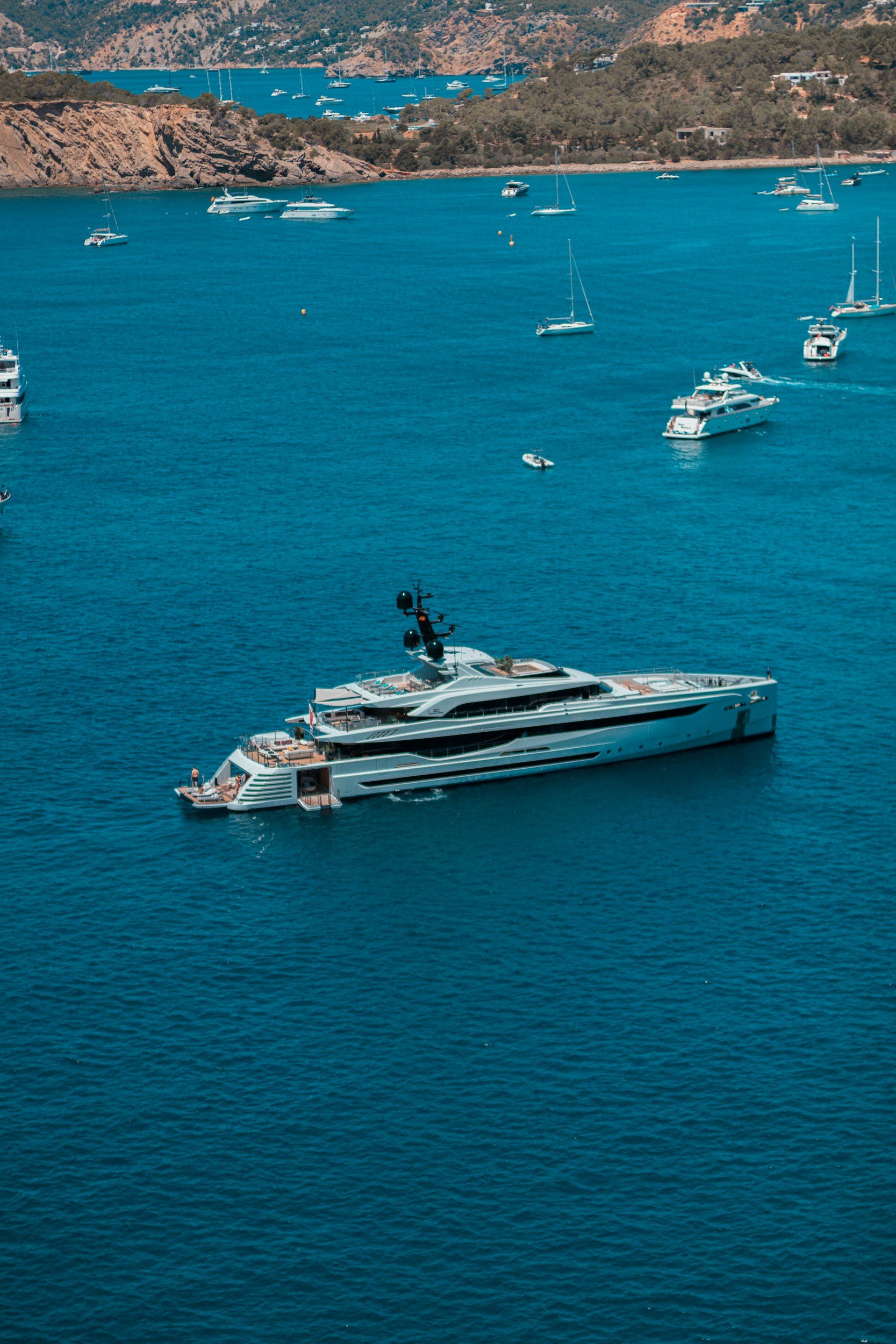 A large white yacht sailing in blue water with several sailboats and motorboats around it, with a green hilly coastline in the background.