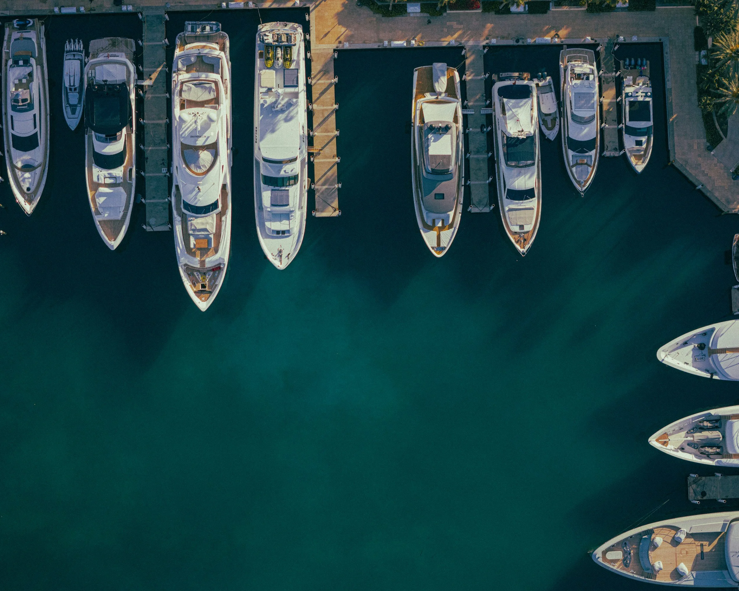 An aerial view of a marina with several yachts docked in calm water and a paved walkway along the edge, with some palm trees.