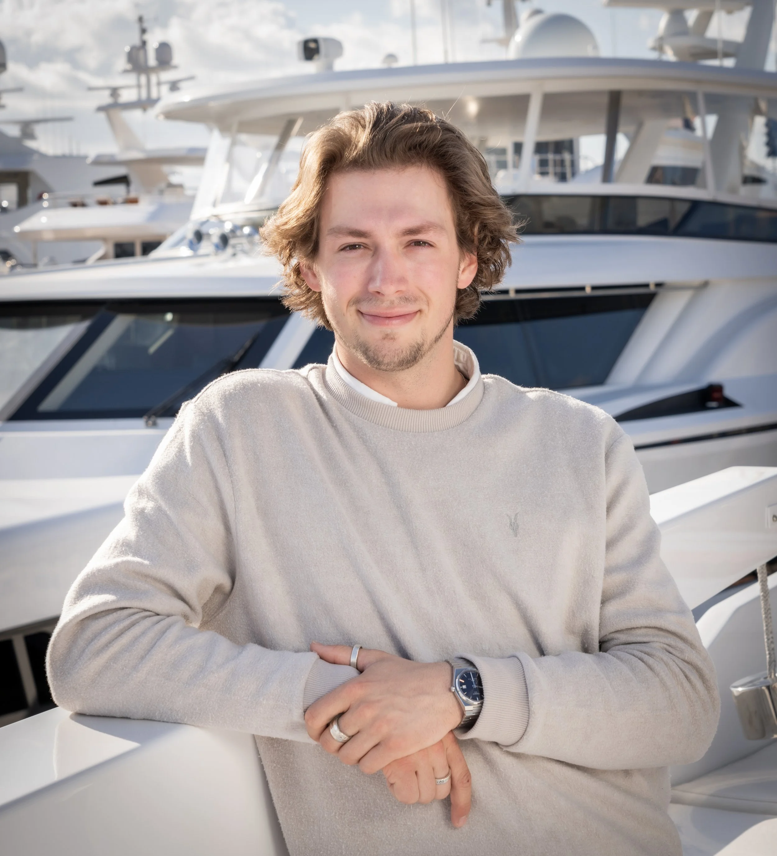 A young man with wavy brown hair, wearing a white sweater and a silver watch, standing in front of yachts docked at a marina.
