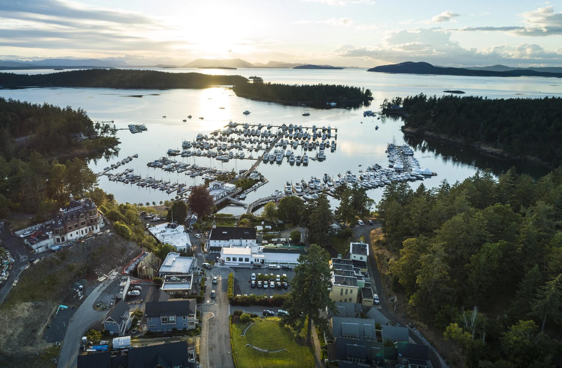 An aerial view of a marina with boats docked, surrounded by lush green trees and residential houses, with a body of water and distant mountains in the background during sunset.