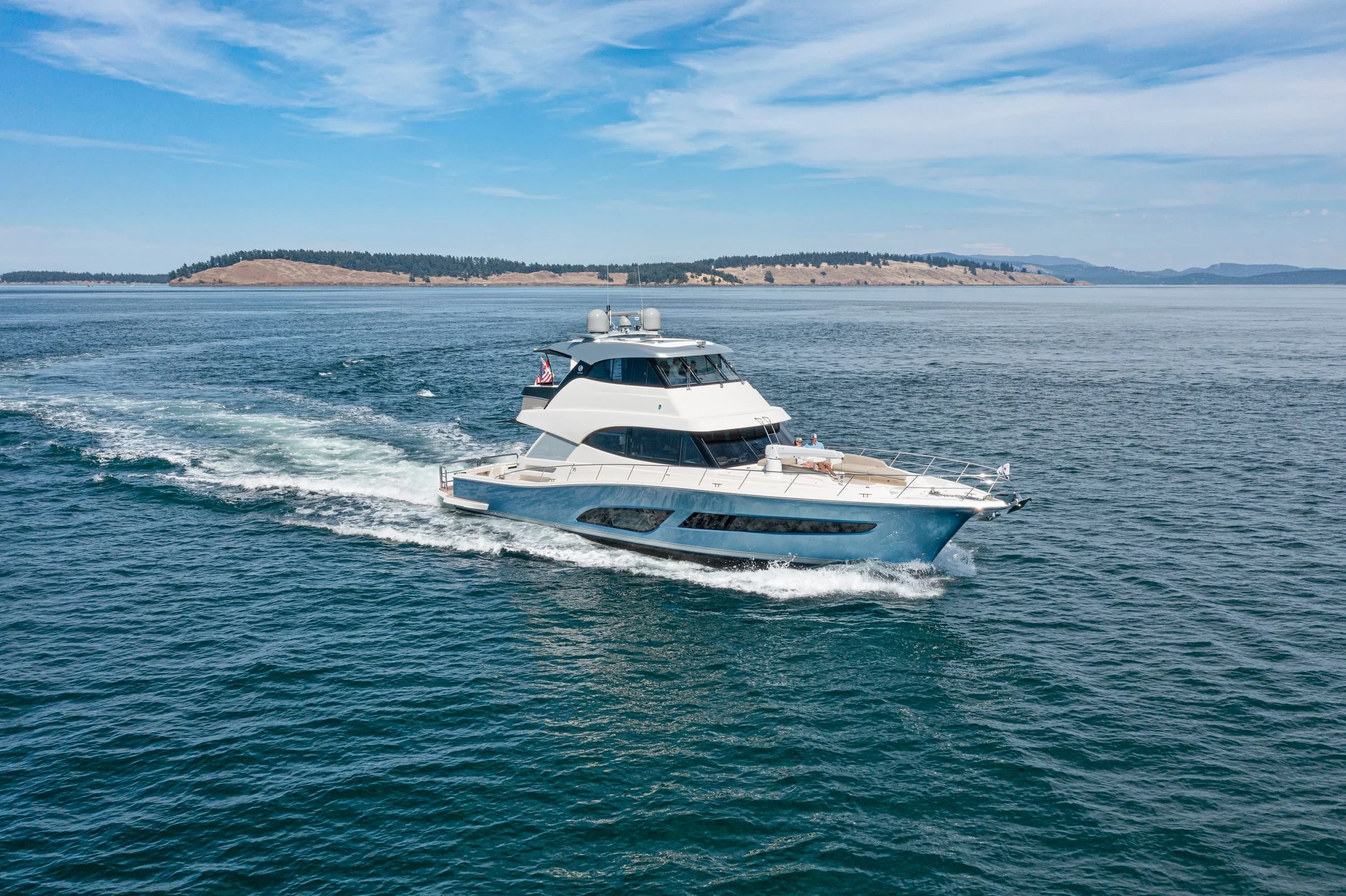 A white yacht cruising on calm blue water with a distant hilly island and partly cloudy sky in the background.