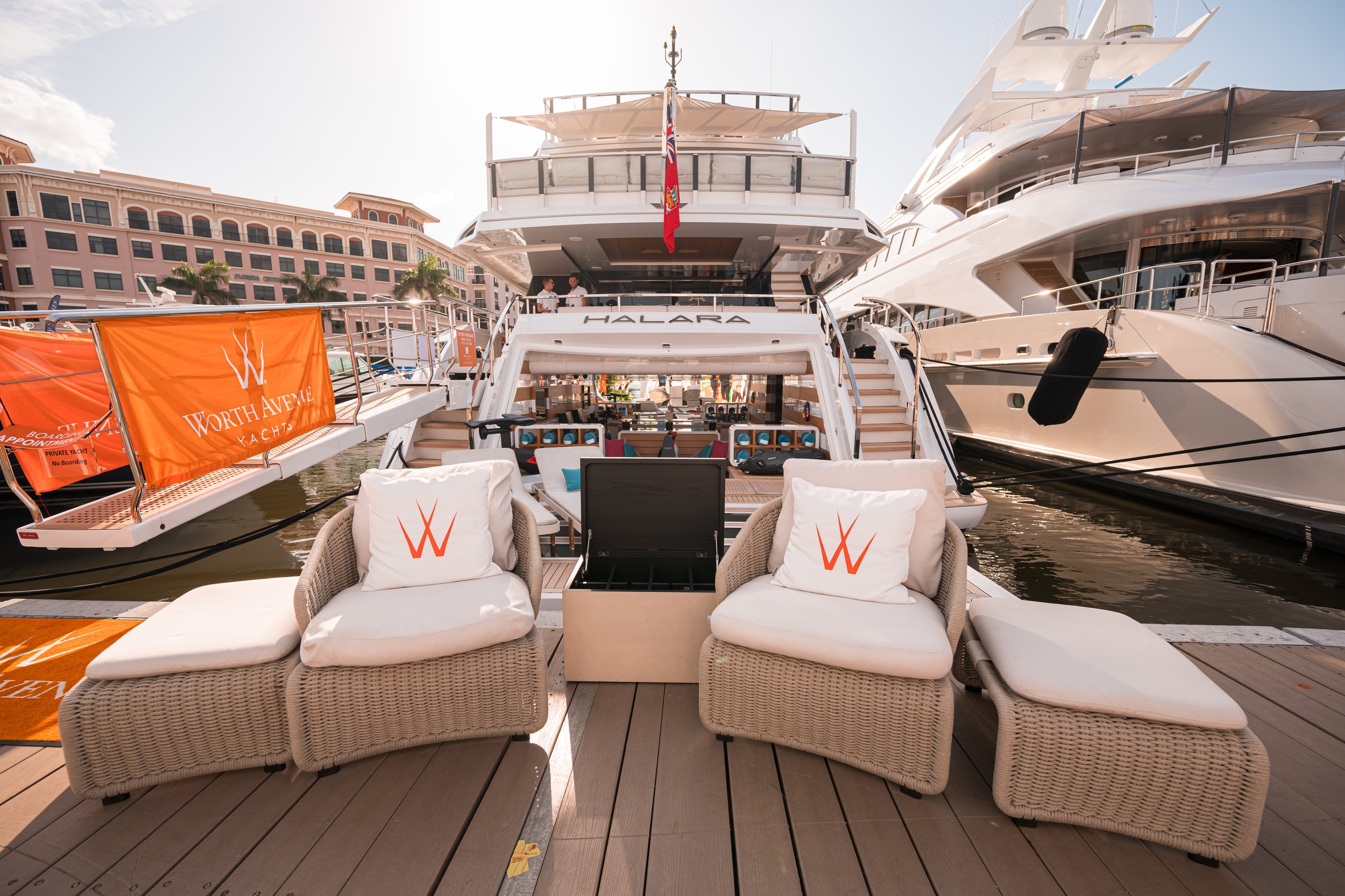 A large luxury yacht docked at a marina, with outdoor seating on the deck, two people talking on the upper level, and a building with pink exterior in the background.