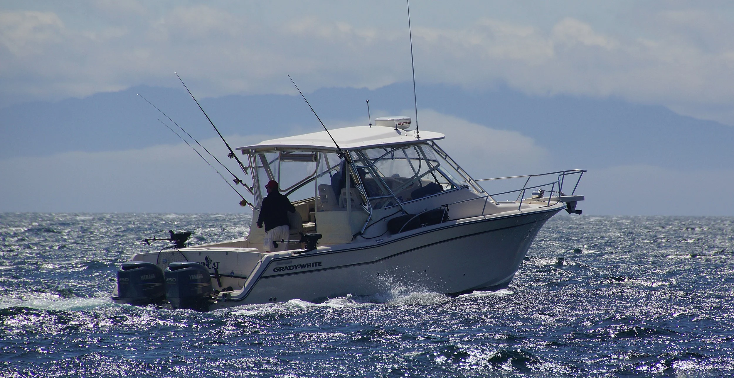 A white Grady-White boat with fishing rods on a cloudy day, moving through the ocean with two Yamaha engines, and a person standing on the deck.