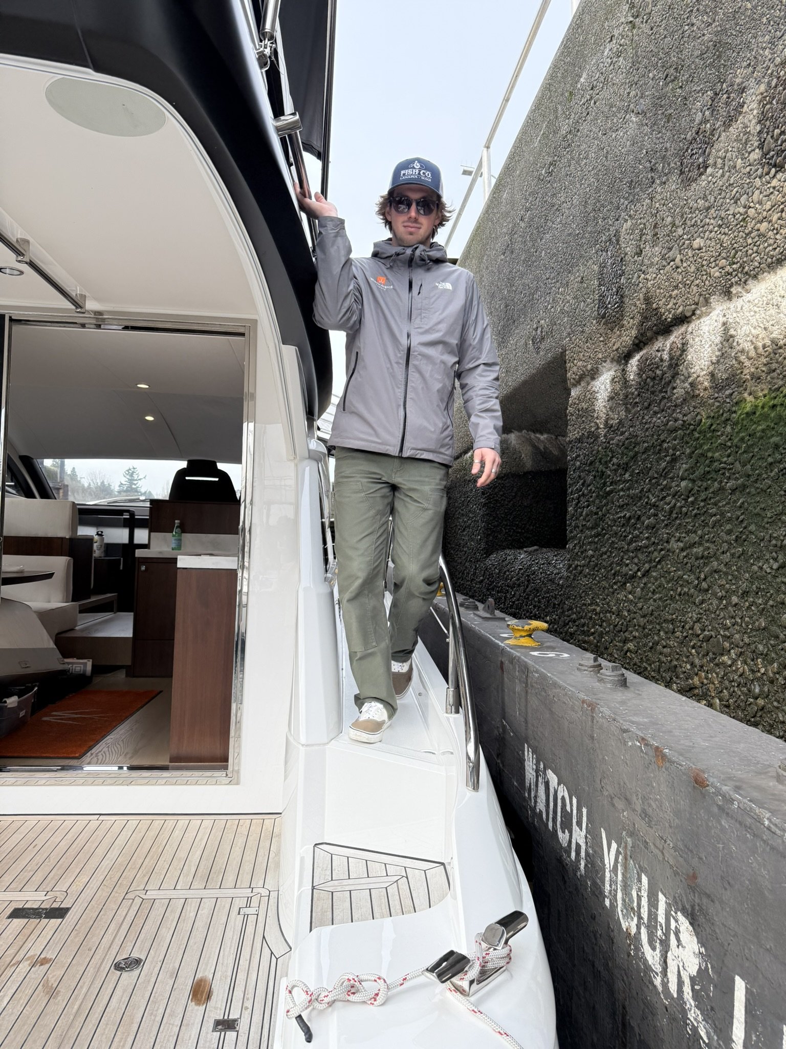 A young man standing on a yacht's deck, holding onto the railing with one hand, next to a large black whale on a truck. The man is wearing sunglasses, a gray jacket, and a baseball cap. The yacht's interior with couches and a wooden table is visible inside.