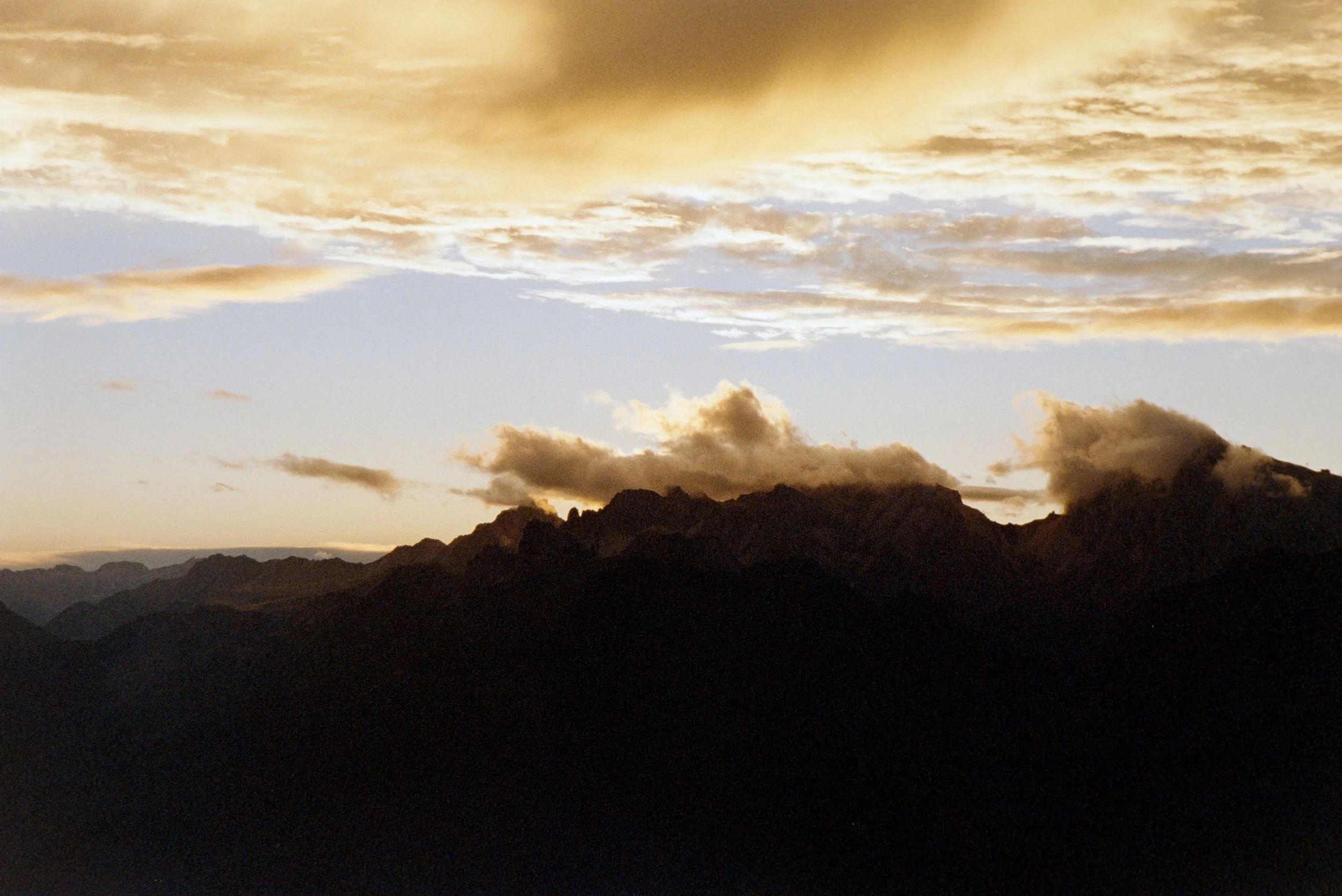 A mountain range silhouettes against a cloudy sky at sunset with golden clouds.