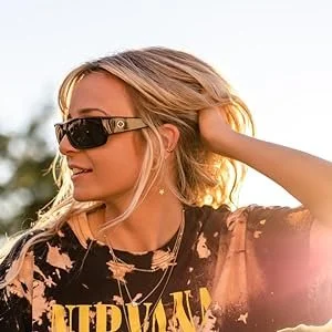A woman with blonde hair wearing sunglasses and a Nirvana T-shirt outdoors, touching her hair.