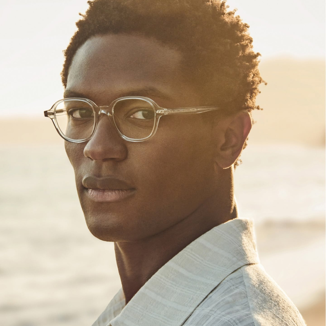 A young man with short, curly hair and glasses looking at the camera with a beach in the background during sunset.