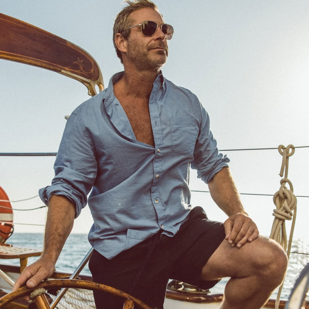 A man with sunglasses, wearing a light blue button-up shirt and black shorts, sitting on a boat with the ocean and sky in the background.