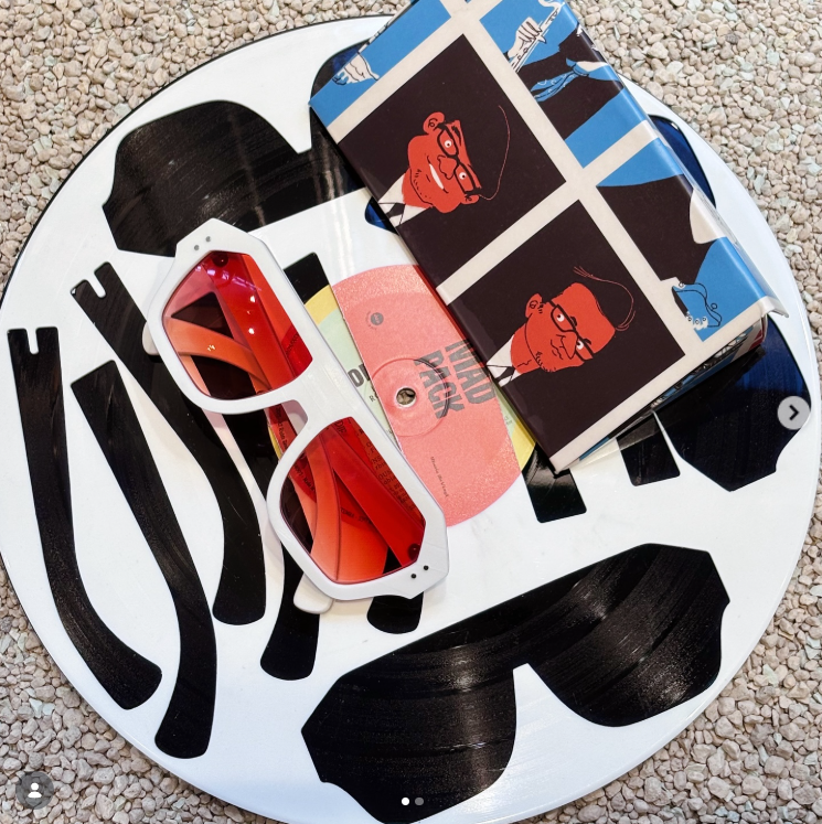 A black and white vinyl record with the words 'The Who' printed on it, a pair of red sunglasses with white frames, and a black and blue box featuring caricatures of a man with glasses and a hat, all placed on a gravel surface.