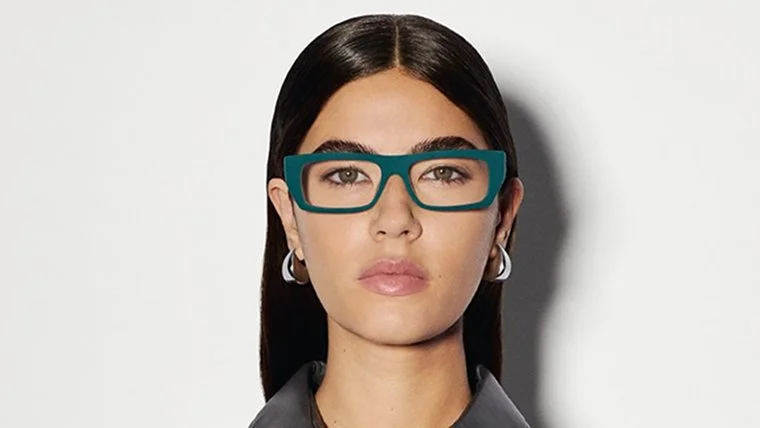 A woman with long dark hair, wearing large teal glasses, hoop earrings, and a gray blazer, against a plain light background.