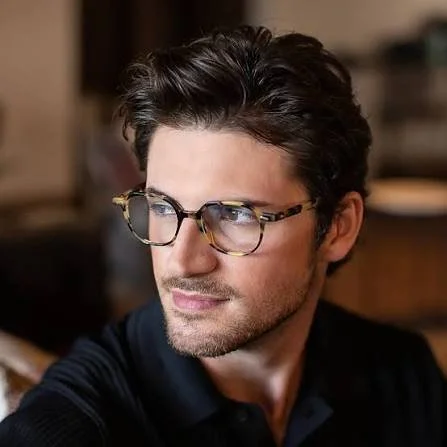 A man with dark hair, wearing glasses, looking to the side, in a room with warm lighting.
