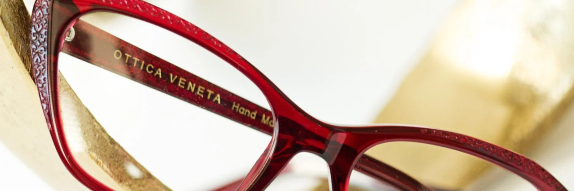Close-up of red eyeglasses with gold lettering on the arm that says 'OTTICA VENETA Hand Made' against a blurred beige background.