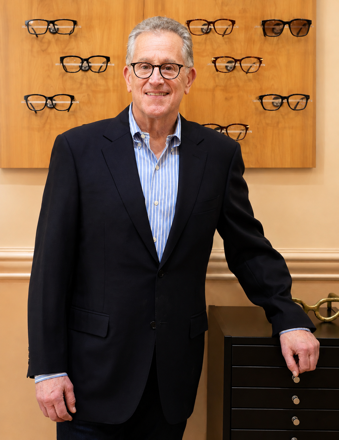 An older man with gray hair and glasses, wearing a black suit and blue striped shirt, stands in an eyewear store in front of a wooden display with multiple pairs of eyeglasses.