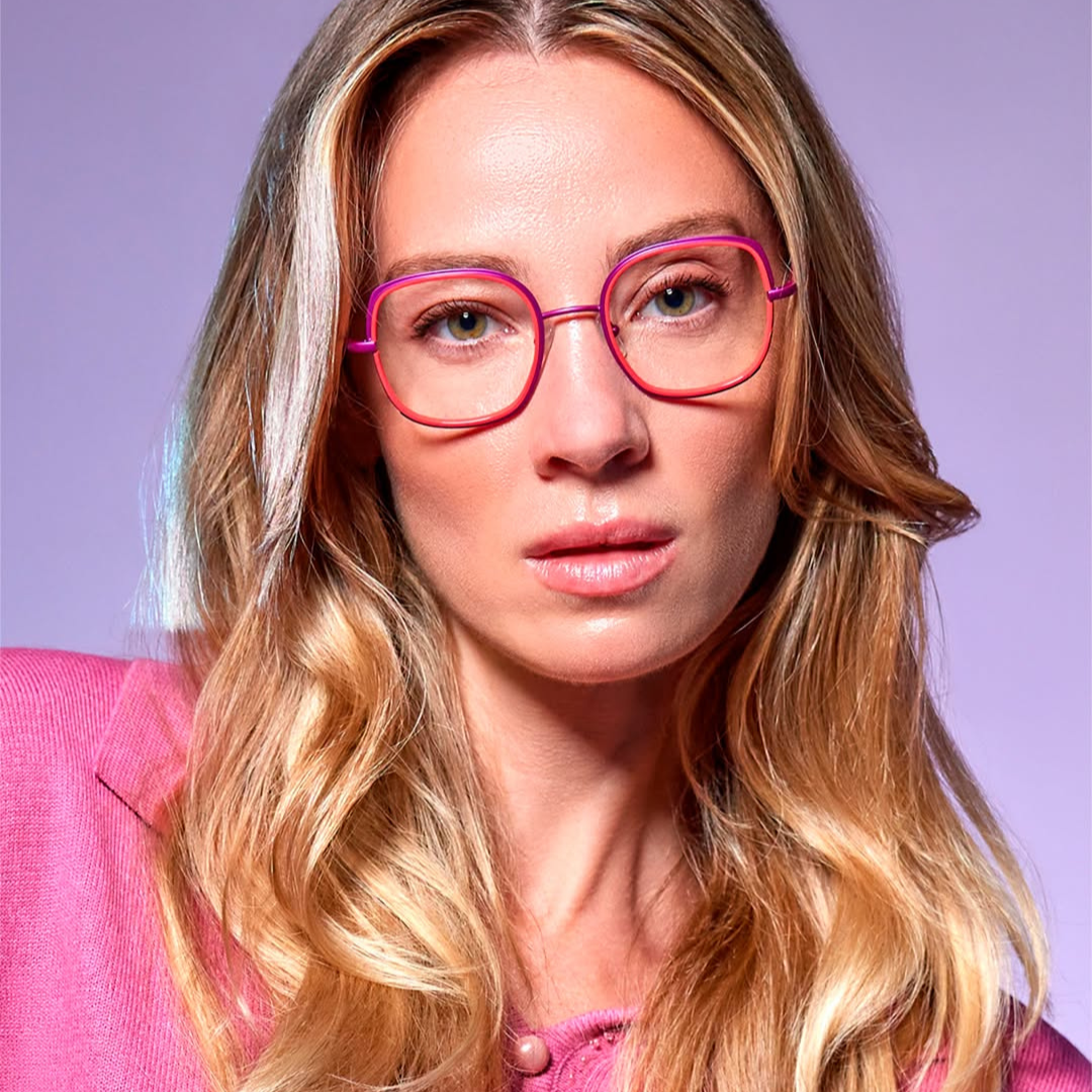 Close-up of a woman with wavy blonde hair wearing pink glasses and a pink blazer against a purple background.