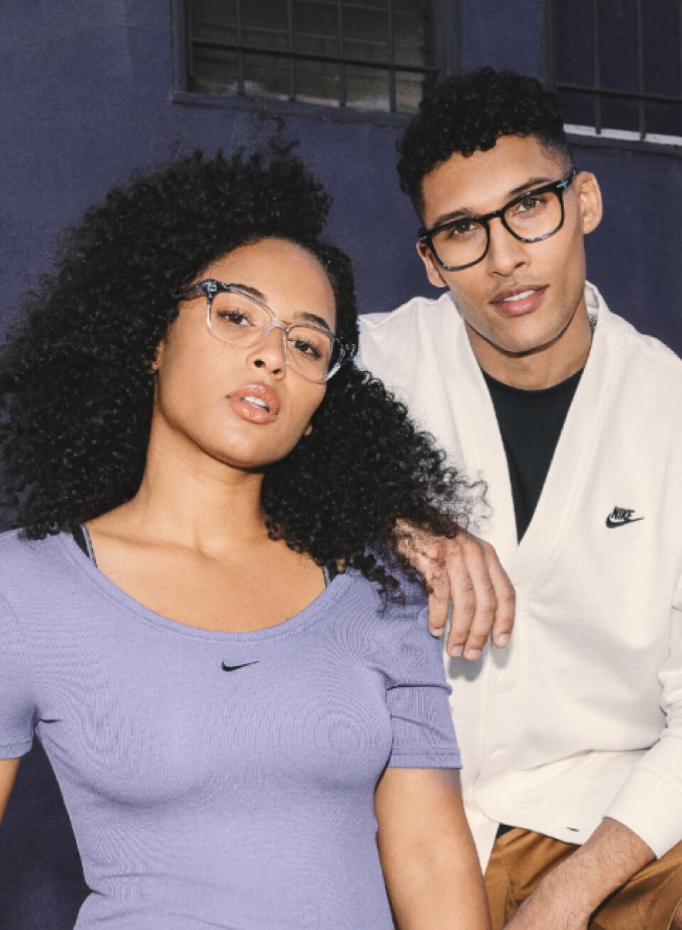 Two young adults, a woman and a man, wearing glasses and casual athletic clothing posing together outside against a dark wall with a window. The woman has curly hair and is wearing a purple Nike shirt, while the man has short curly hair and is wearing a white Nike hoodie.