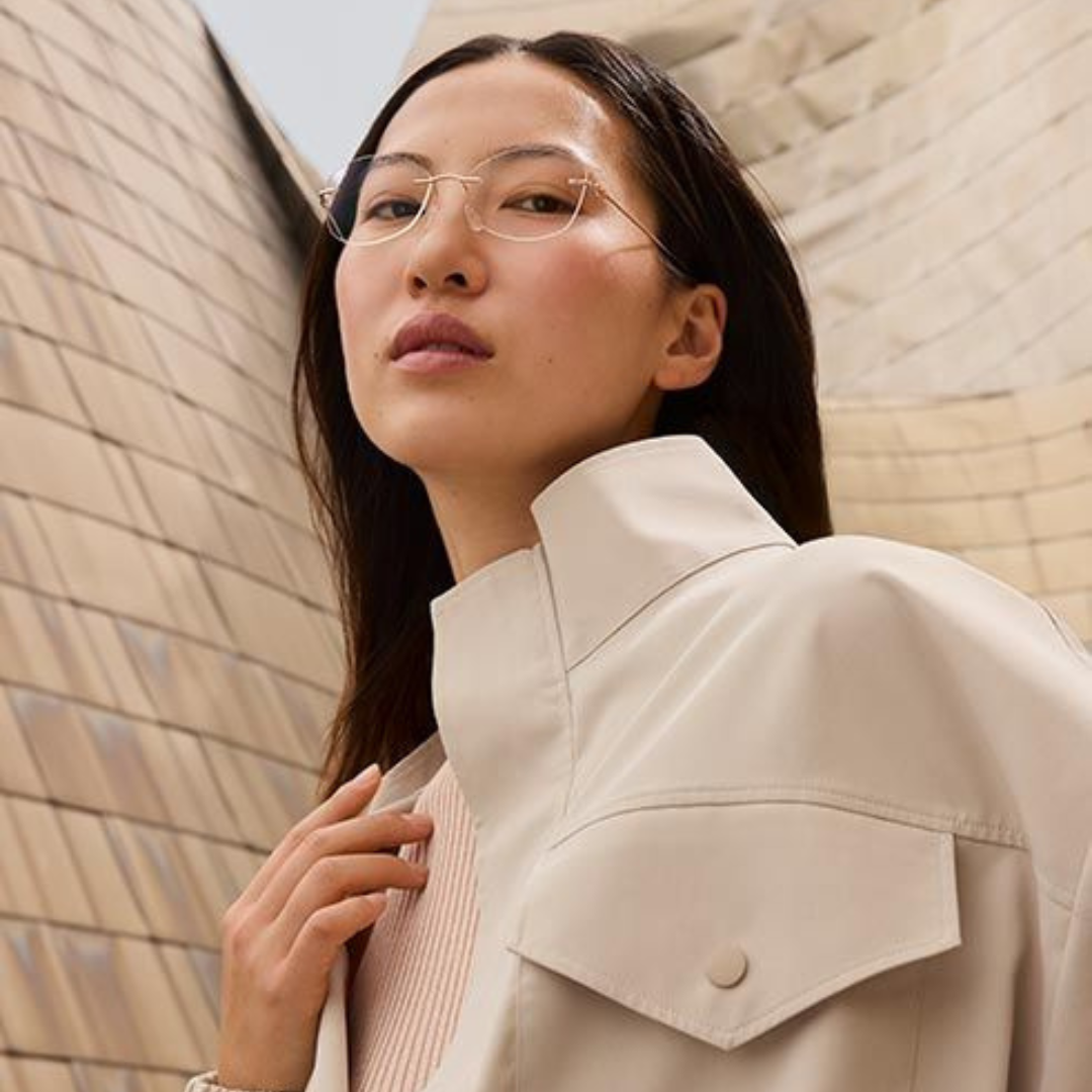 A woman with long dark hair, wearing glasses and a beige outfit, standing against a brick wall.