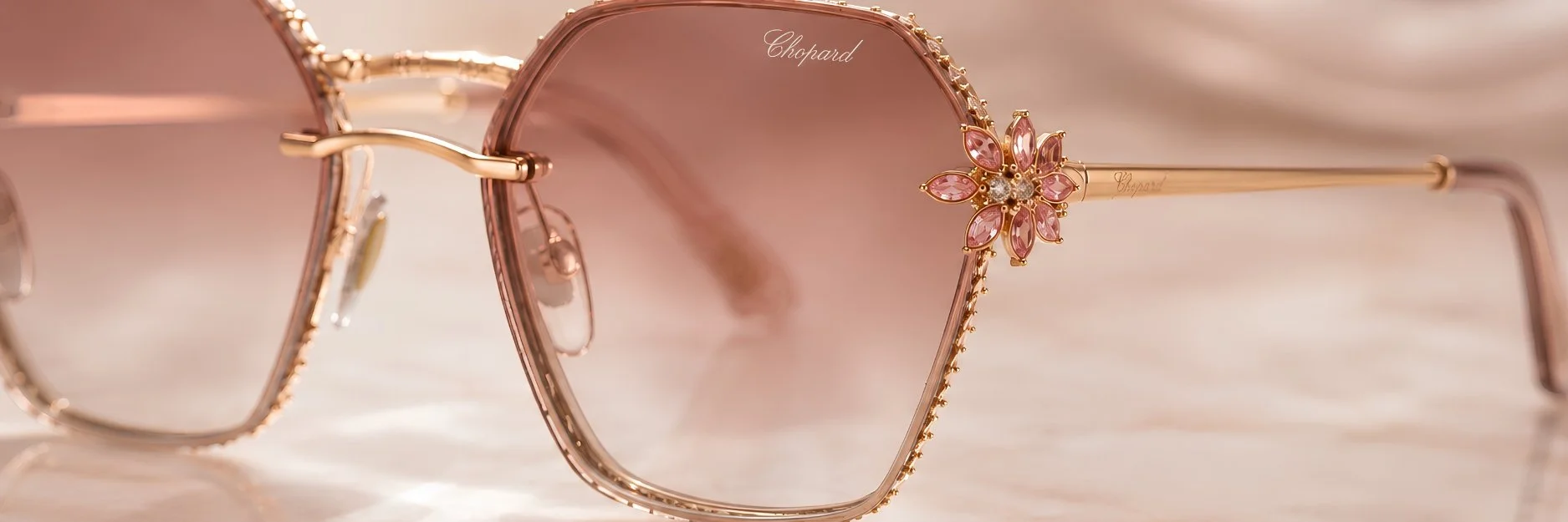 Close-up of a pair of pink-tinted sunglasses with gold frames and decorative pink gemstone flower accents on the arms, resting on a shiny surface.
