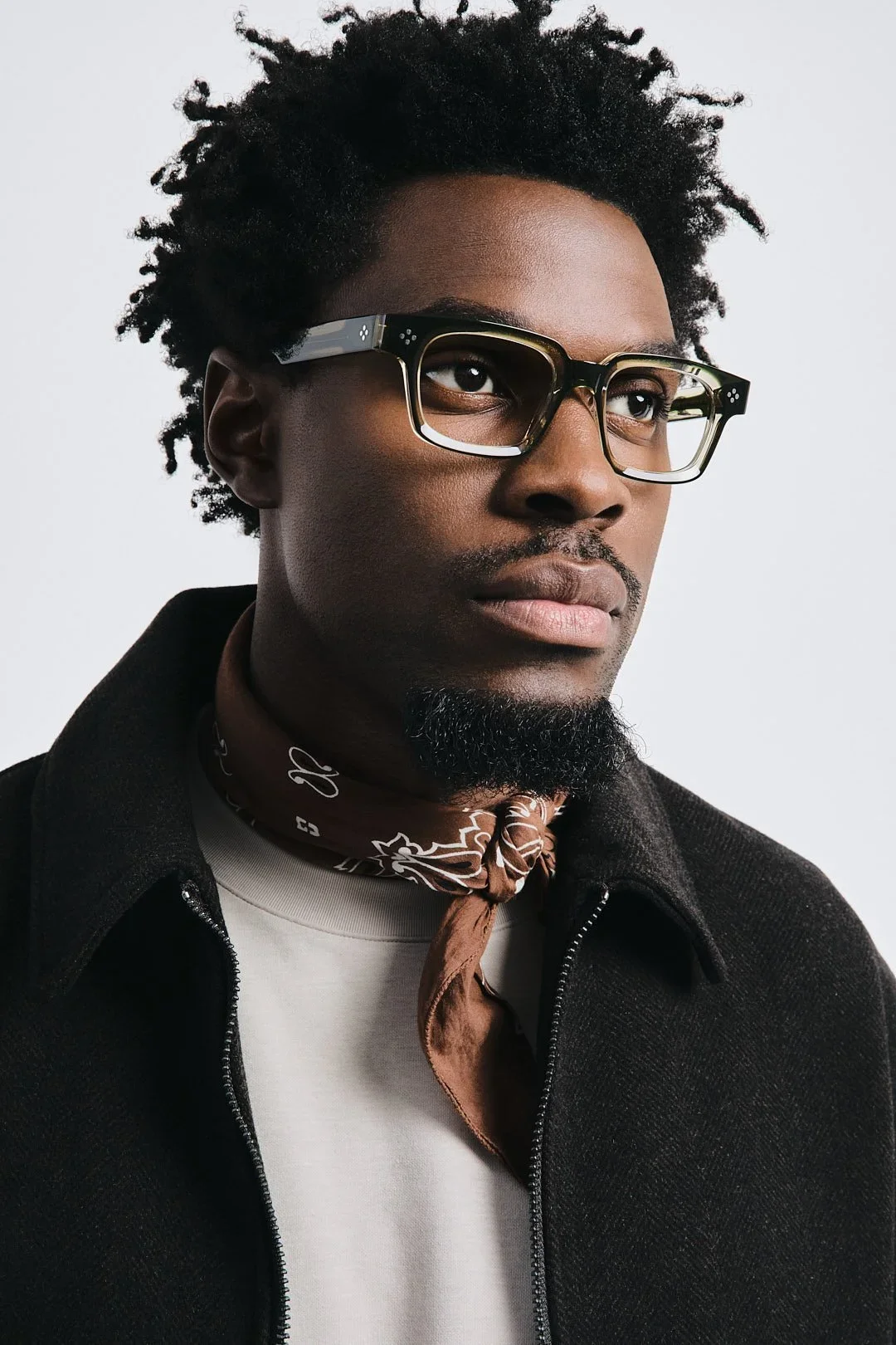 A man with dark skin, wearing large black glasses, a brown patterned bandana around his neck, a white shirt, and a black zip-up jacket, with short curly hair and a beard, against a plain white background.