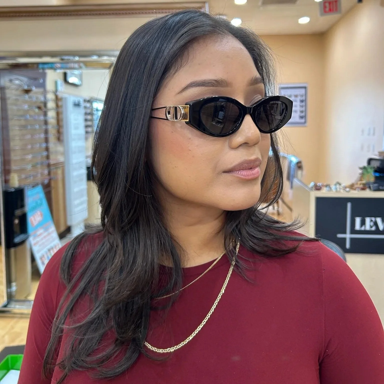 A woman with dark hair wearing black designer sunglasses and a gold chain necklace, standing indoors with store shelves and a sign visible in the background.