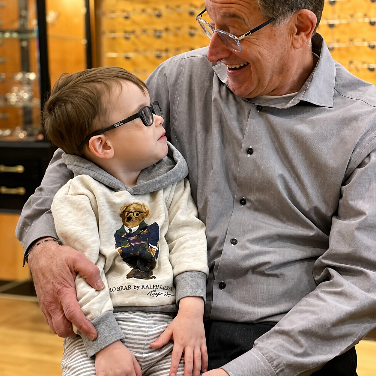 An elderly man smiling and looking at a young boy, who is sitting on his lap, inside a store with glass display cases in the background.