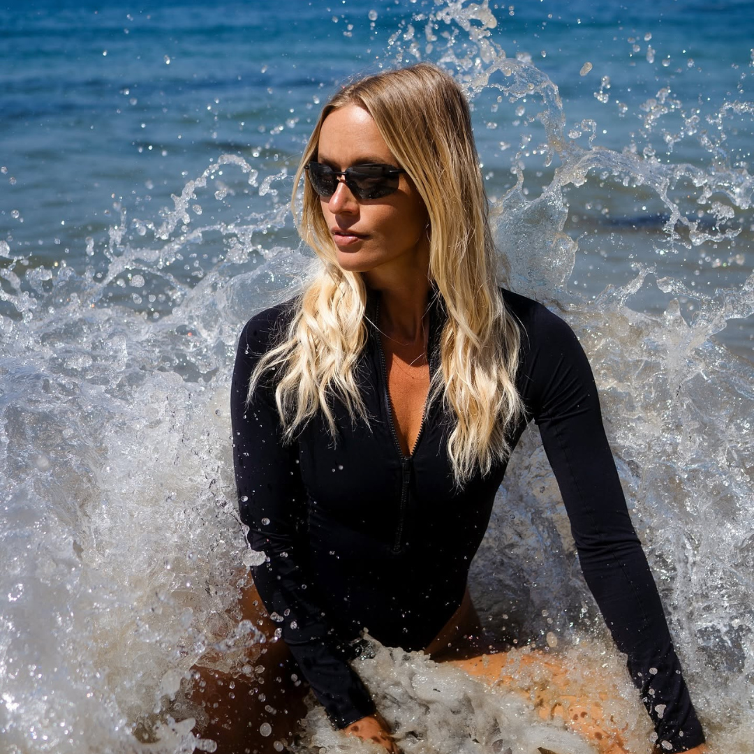 Woman with long blonde hair wearing sunglasses and a black wetsuit sitting in the ocean with splashing waves around her.