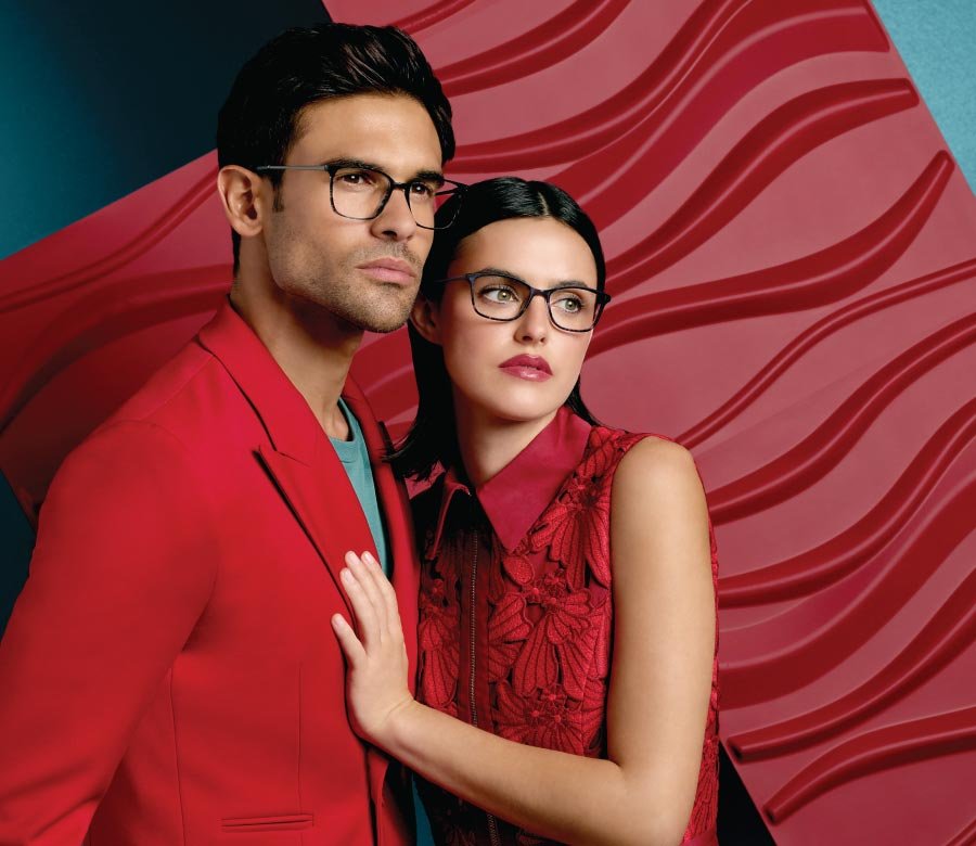 A man and a woman wearing glasses and red clothing, standing close together in front of an abstract red and teal background.