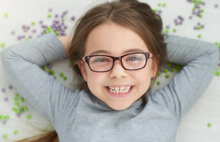 Smiling girl with glasses lying on her back on a white surface sprinkled with green and purple beads.