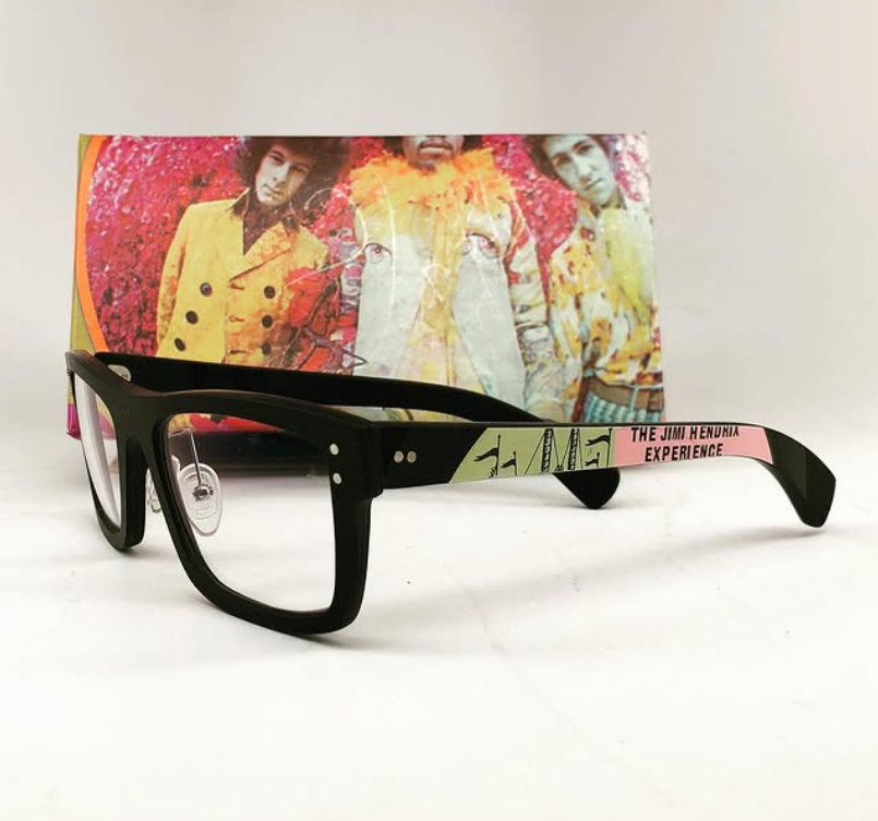 A pair of black glasses with a decorative pink and green arm resting on a white surface in front of a colorful album cover featuring images of Jimi Hendrix in bright yellow and floral clothing.