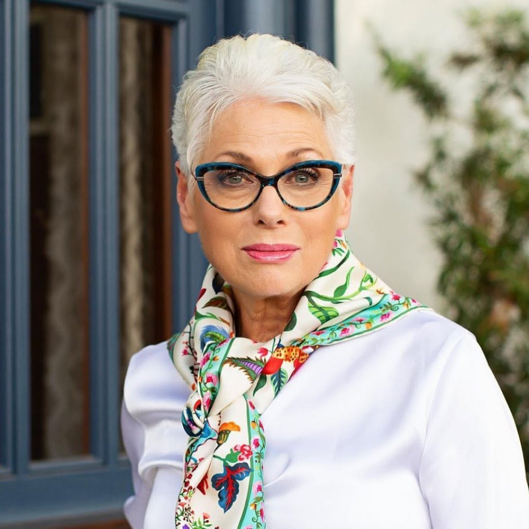 An older woman with short, silver hair and glasses, wearing a white shirt and a colorful scarf, standing outdoors with a blurred background.