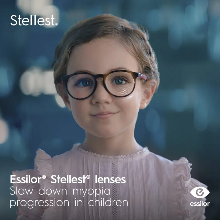 A young girl wearing glasses and a light pink blouse, smiling at the camera, with the logo 'Stellest' in the top left corner and text about Essilor Stellest lenses and myopia progression.