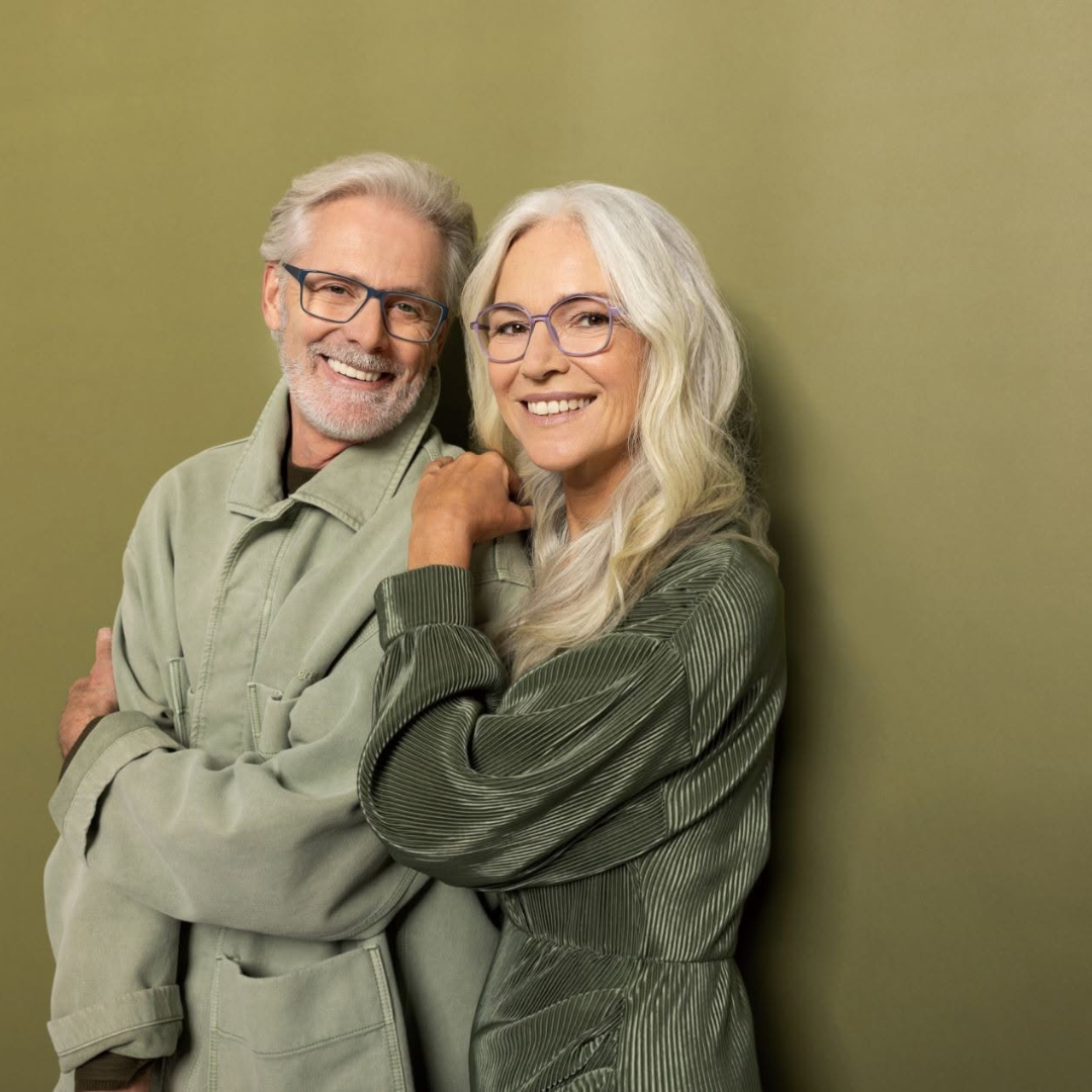 A smiling elderly couple with glasses, standing close together against a green wall.