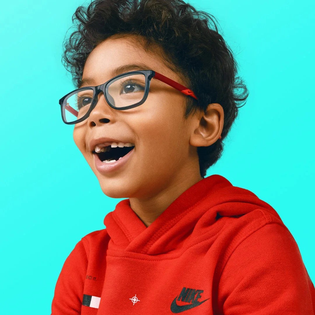 A young boy with curly dark hair wearing glasses and a red Nike hoodie, smiling and looking off to the side against a light blue background.