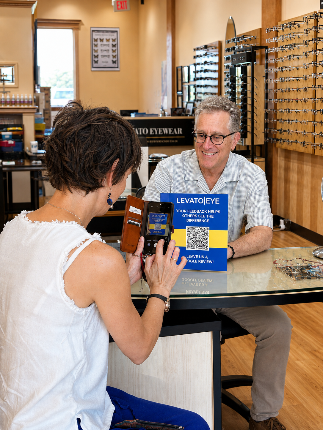 Customer and employee at an eyewear store with eyeglasses display in the background, customer being recorded on a smartphone, and a sign promoting reviews on the counter.