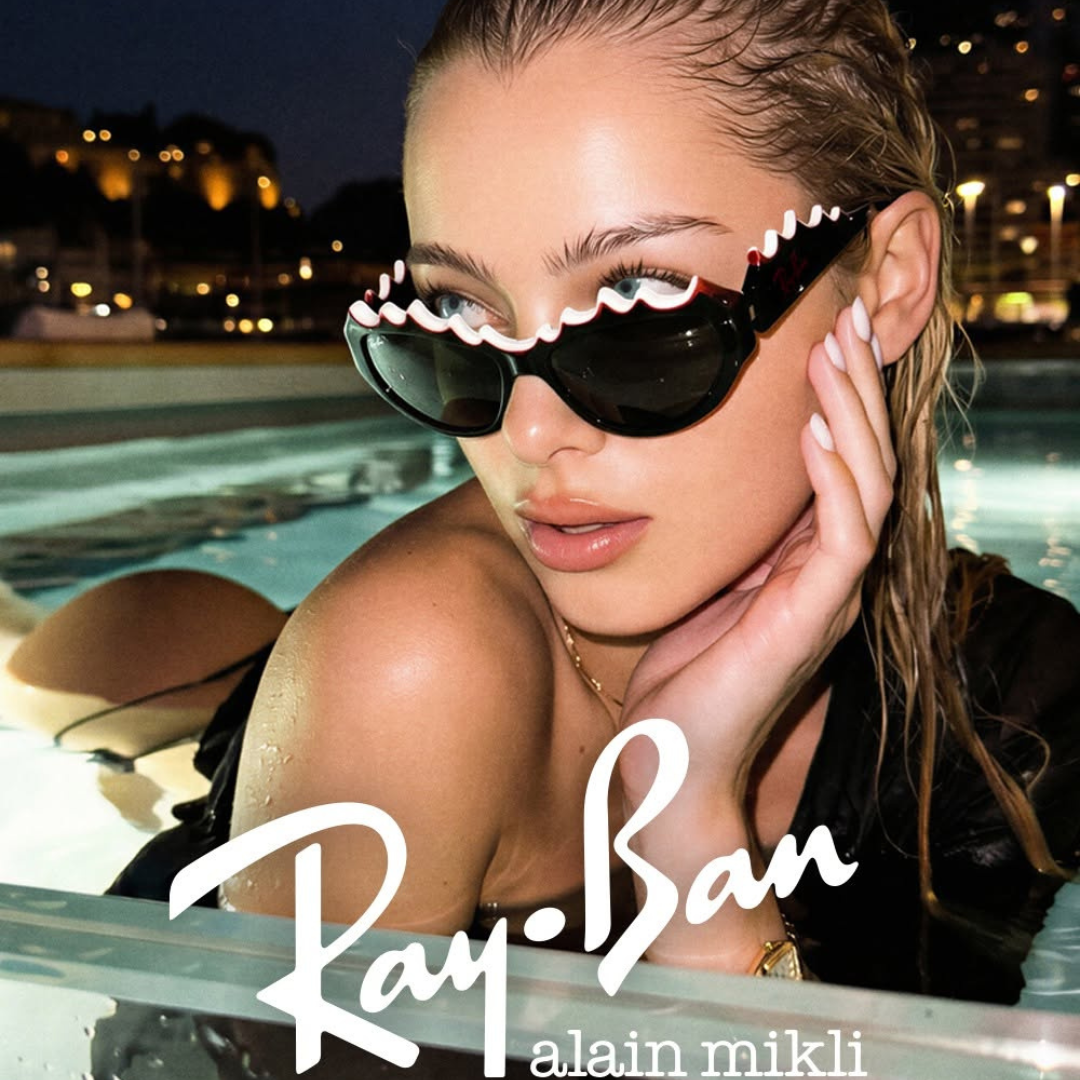 Close-up of a woman with wet hair wearing black sunglasses with red and white details, resting her hand on her face while partially submerged in a swimming pool at night, with city lights in the background, and the Ray-Ban logo and the name 'Alain Mikli' visible.