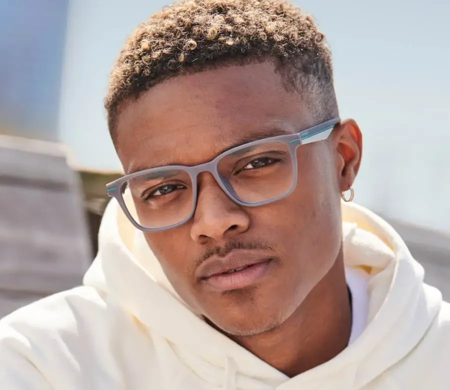 Close-up of a young man with curly hair, wearing light blue glasses, a white hoodie, and an earring, outdoors on a sunny day.