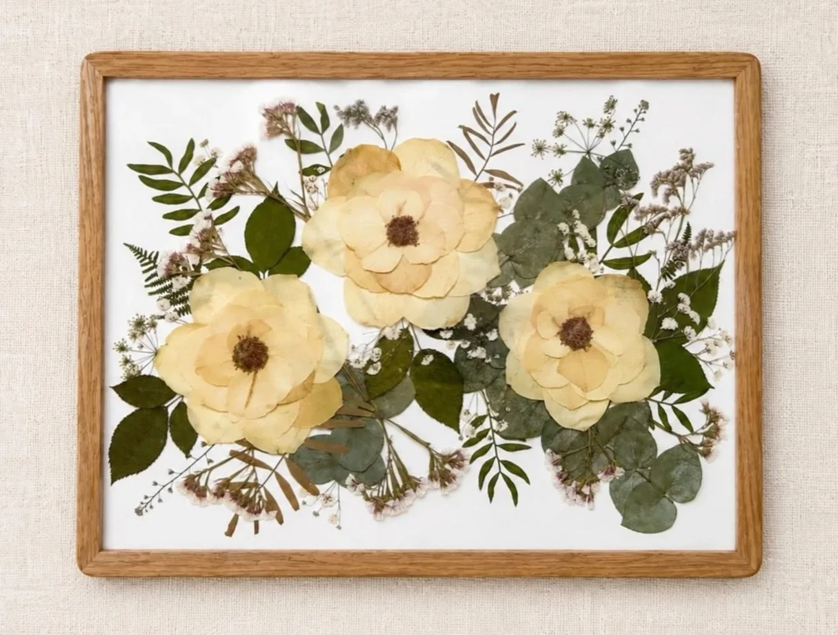 Framed artwork of dried pressed flowers and leaves on a white background.