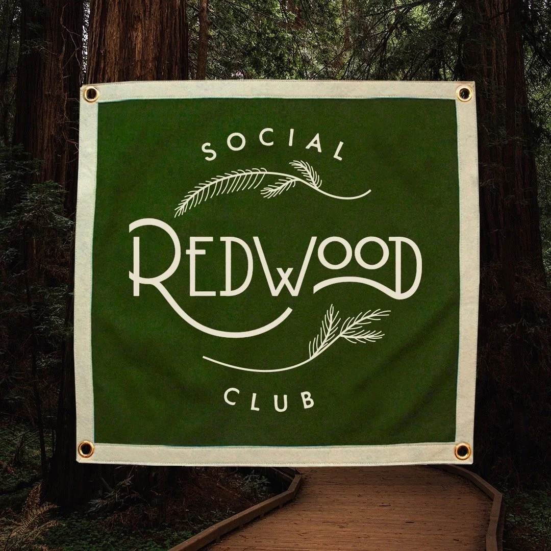 A green sign with white lettering that reads "Social Red Wood Club" and decorative leaf designs, mounted on a wooden post in a forest setting.