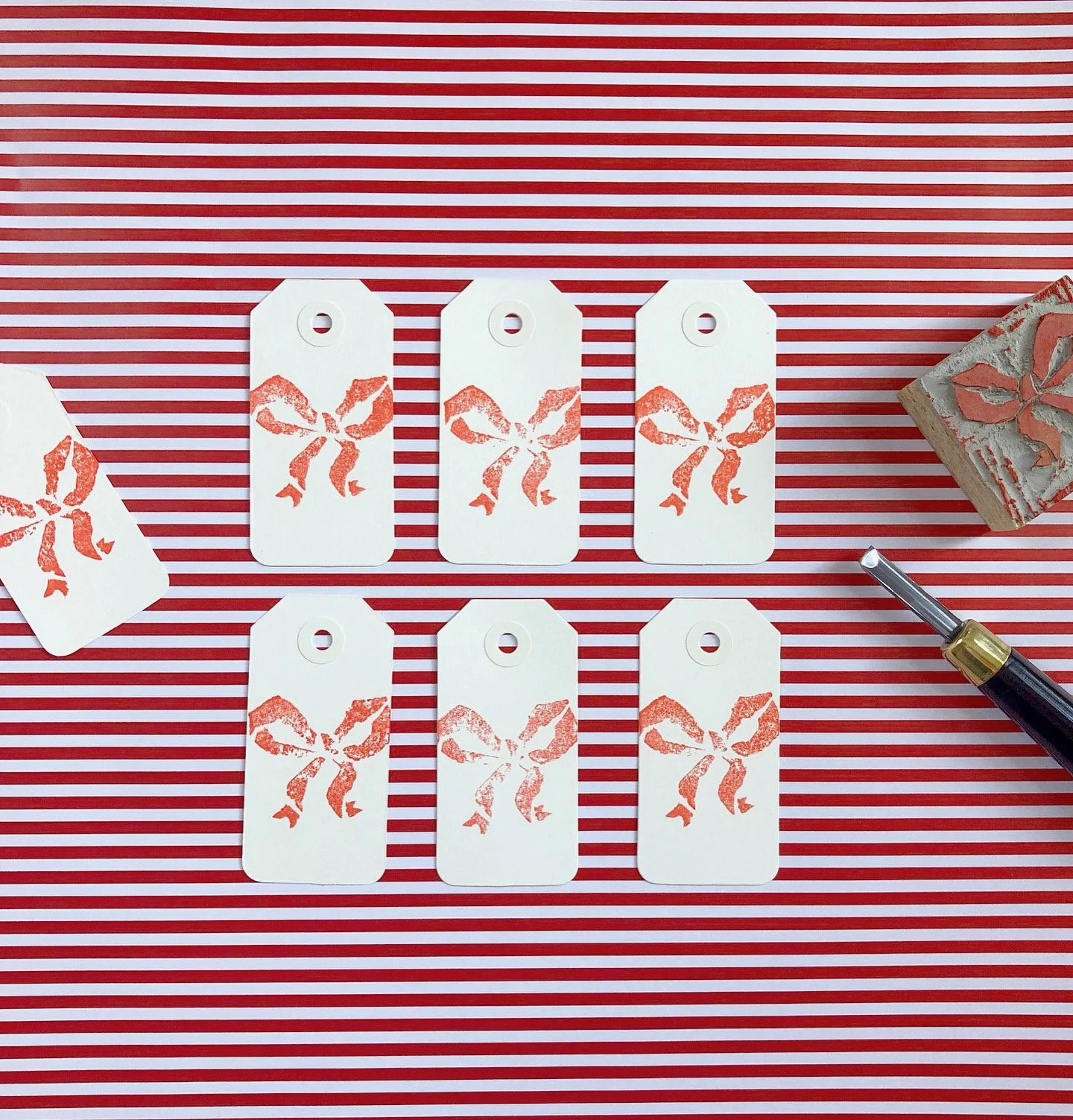 Six white gift tags with a red bow design printed on them, arranged on a red and white striped background. A hole punch at the top of each tag. Part of a rubber stamp with a red bow design and a metallic pen on the right side.