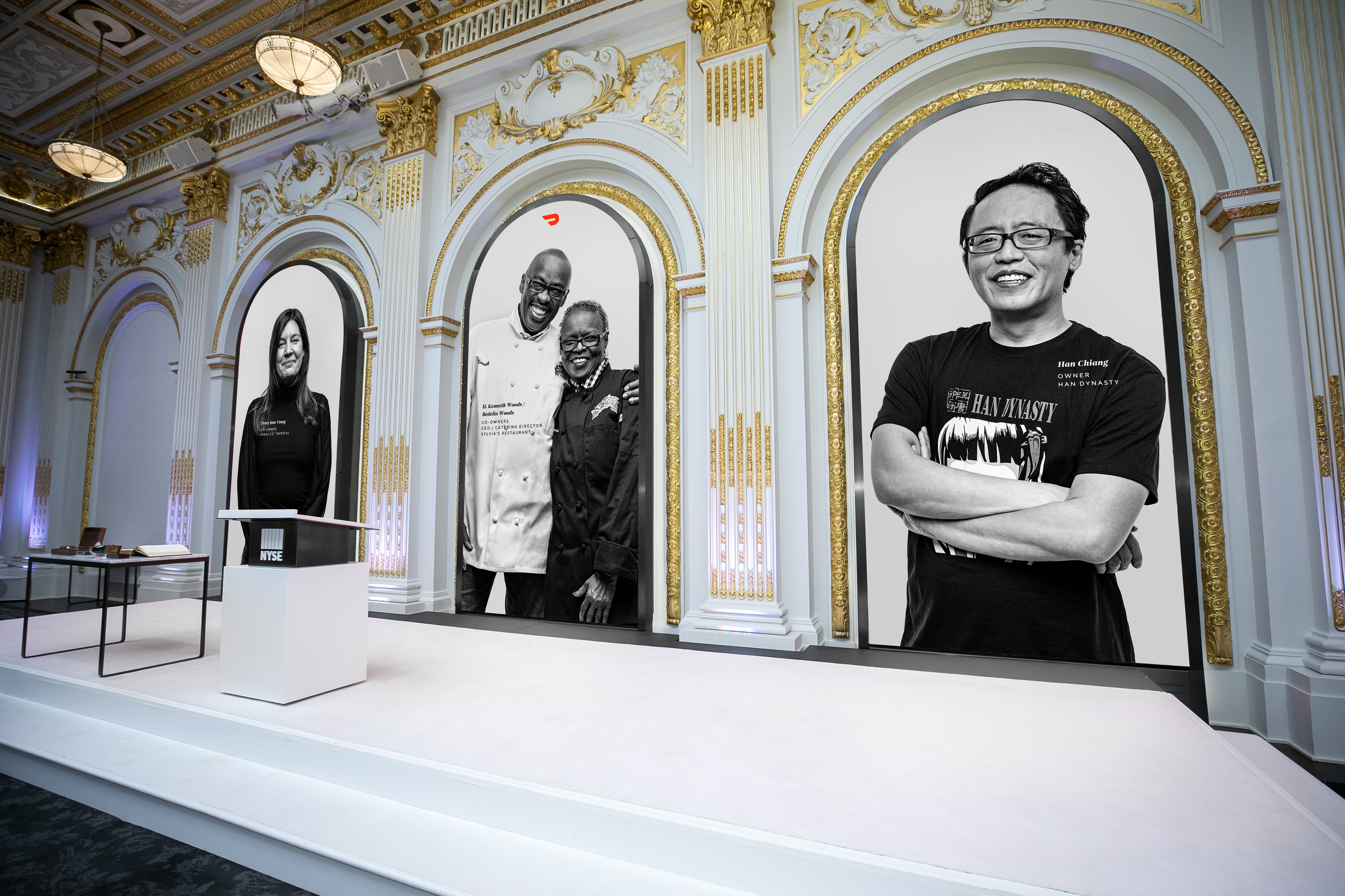 Four people standing on a stage with elaborate white and gold decor. Two large framed black-and-white photos are in the background, one of a woman and the other of two men, including a chef and a man in a black t-shirt. The stage has a white podium w