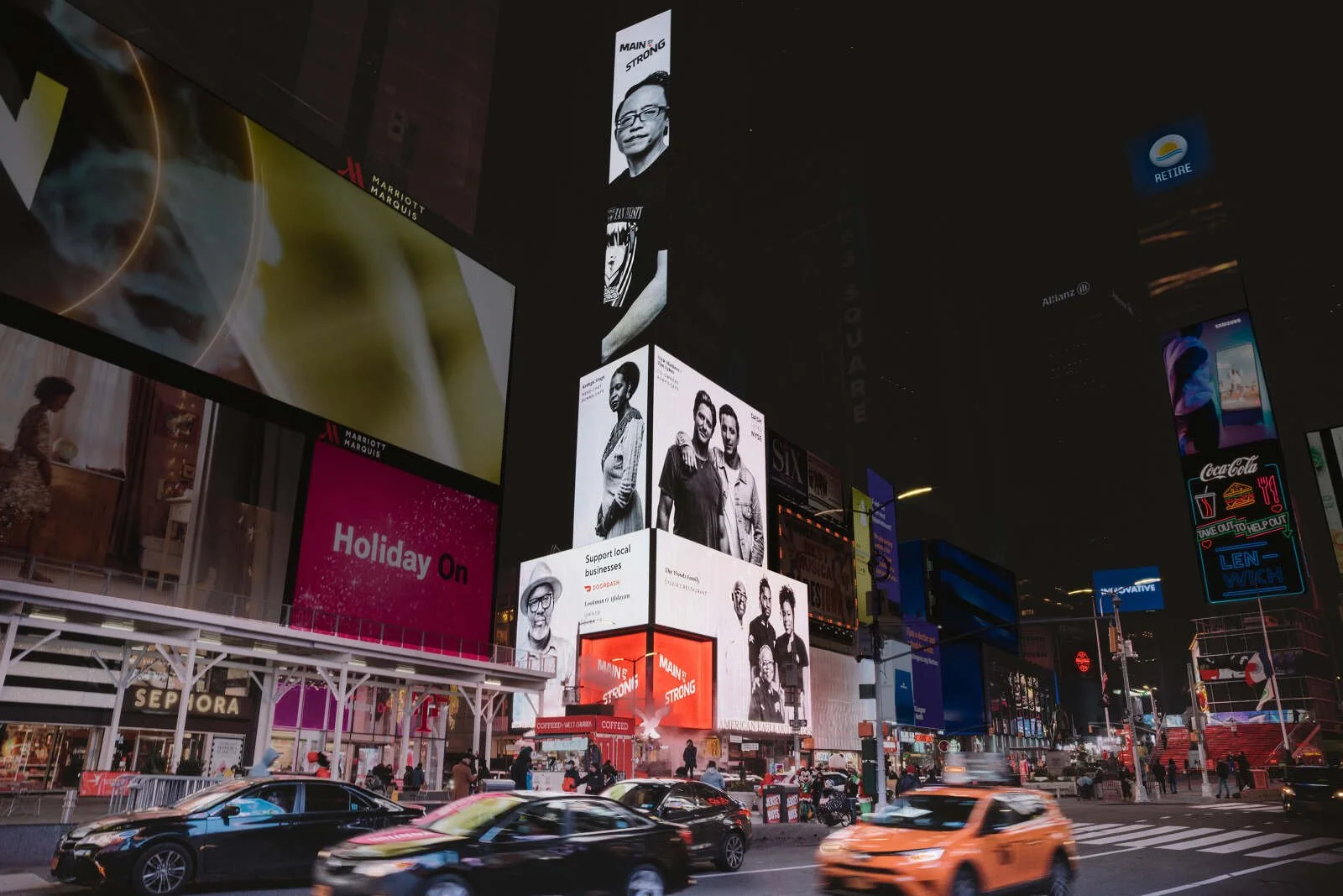 Nighttime view of Times Square in New York City with illuminated digital billboards, including advertisements for Marriott Marquis, Coca-Cola, and various brands. Cars are moving on the street, and people are walking on the sidewalks.