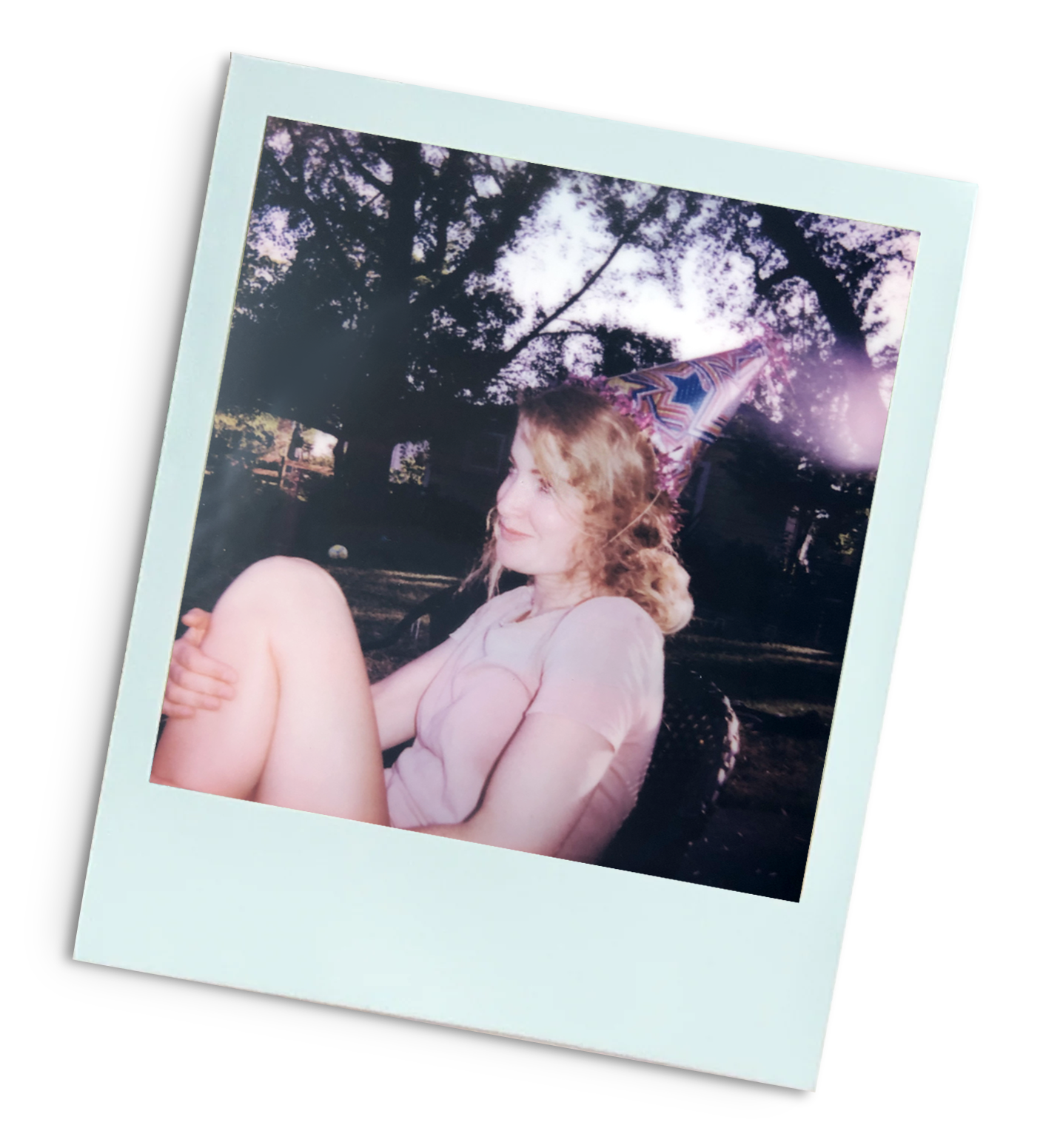 A woman with curly hair wearing a pink shirt and a colorful party hat sits outdoors, smiling with her eyes closed, holding her knee. The background includes trees and a fence, captured during sunset or dusk.